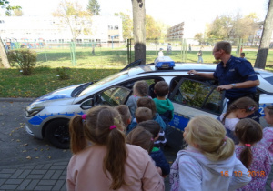 Dzieci stoją z policjantami przy radiowozie policyjnym. Policjant otwiera drzwi auta.